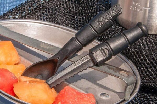 Two black Ka-Bar utensil tools, a spork and a serrated knife, rest on a metal camping dish filled with chunks of fruit, with a textured background and a stainless steel container nearby.