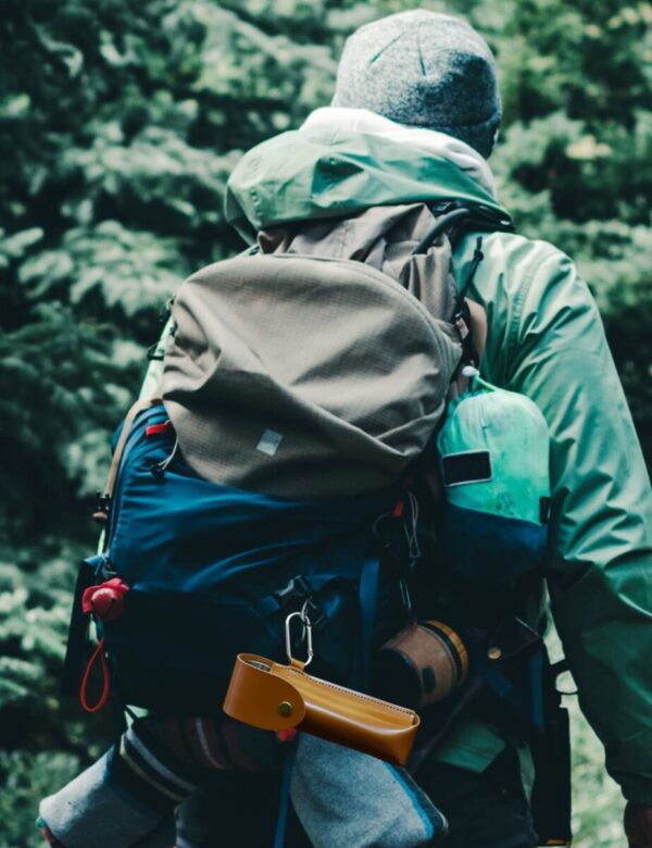 A person wearing a green jacket and a gray beanie is hiking through a forest with a large backpack. Various outdoor gear, including a water bottle and pouches, are attached to the backpack.